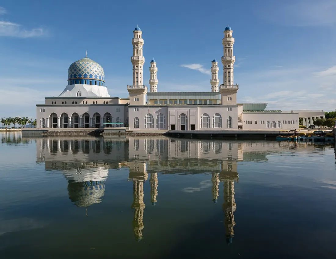 A white mosque with a blue patterned dome and four minarets, mirrored in still water, in Kota Kinabalu.