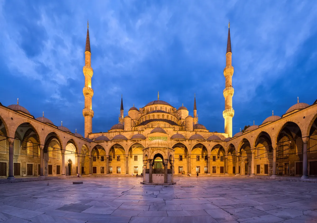 The courtyard of the Sultan Ahmed Mosque in Istanbul at dusk, its domes and minarets lit against a deep blue sky.