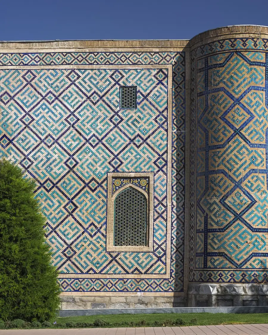 A wall of blue and turquoise geometric tilework with a small latticed window, at the Sher-Dor Madrasa in Samarkand.